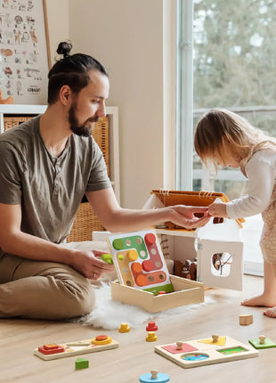 A man and a young girl play on a wooden floor with colorful educational toys, including the Tiny Land® Montessori Complete Baby Set (0-24 months), encouraging sensory play. The bright room has large windows and wicker baskets in the background.