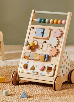 The Tiny Land® Versatile Natural Wooden Baby Walker, featuring colorful beads, gears, shapes, polka dot sides, and four wheels, sits on a woven rug with scattered wooden blocks—perfect for sensory development.