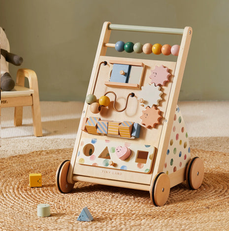 The Tiny Land® Versatile Natural Wooden Baby Walker, featuring colorful beads, gears, shapes, polka dot sides, and four wheels, sits on a woven rug with scattered wooden blocks—perfect for sensory development.