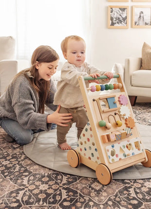 A woman smiles as she kneels on a rug, supporting a toddler who pushes the Tiny Land® Versatile Natural Wooden Baby Walker in a bright, cozy living room, encouraging the child's fine motor skill development.