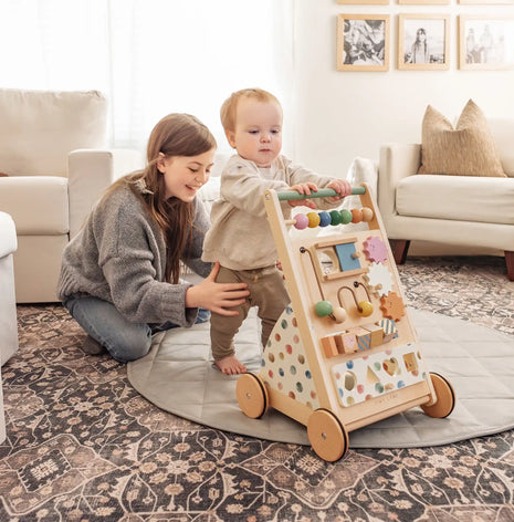 A woman smiles as she kneels on a rug, supporting a toddler who pushes the Tiny Land® Versatile Natural Wooden Baby Walker in a bright, cozy living room, encouraging the child's fine motor skill development.