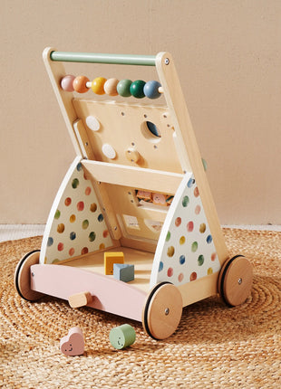 The Tiny Land® Versatile Natural Wooden Baby Walker, with polka dot sides, colorful abacus beads, and shape sorter holes, sits on a woven rug to encourage sensory development. Geometric shapes are scattered nearby.