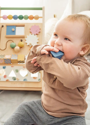 A baby sits on a mat, chewing on a blue block, with the Tiny Land® Versatile Natural Wooden Baby Walker in the background to encourage sensory development.