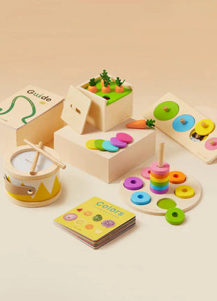 The Tiny Land® Montessori Complete Baby Set (0-24 months) features a drum, stacking rings, carrot harvest game, matching disks, and a color book on a beige surface with a guide booklet to support sensory play and fine motor skills.