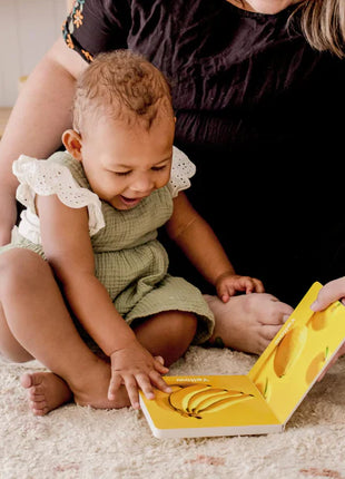 A smiling baby in a green outfit explores the Tiny Land® Montessori Complete Baby Set (0-24 months) on a rug with an adult in a black dress, enjoying sensory play with the colorful picture book’s yellow pages.