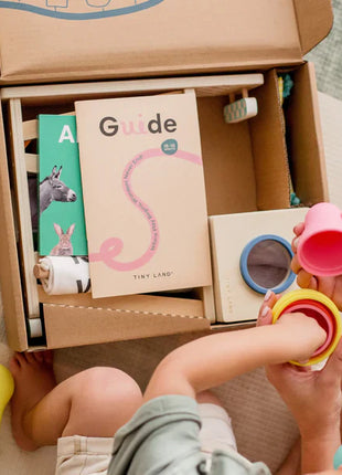 A child and adult enjoy the Tiny Land® Montessori Complete Baby Set (0-24 months), featuring educational toys, colorful stacking cups for motor skills, a Guide booklet, and illustrated animal cards on a patterned mat.