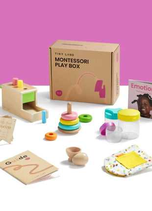 The Tiny Land® Montessori Complete Baby Set (0-24 months) features stacking rings, wooden toys, jars, cards, a guidebook, cloth, and the book Emotions—ideal for sensory play and motor skill development—shown on a white surface with a pink background.